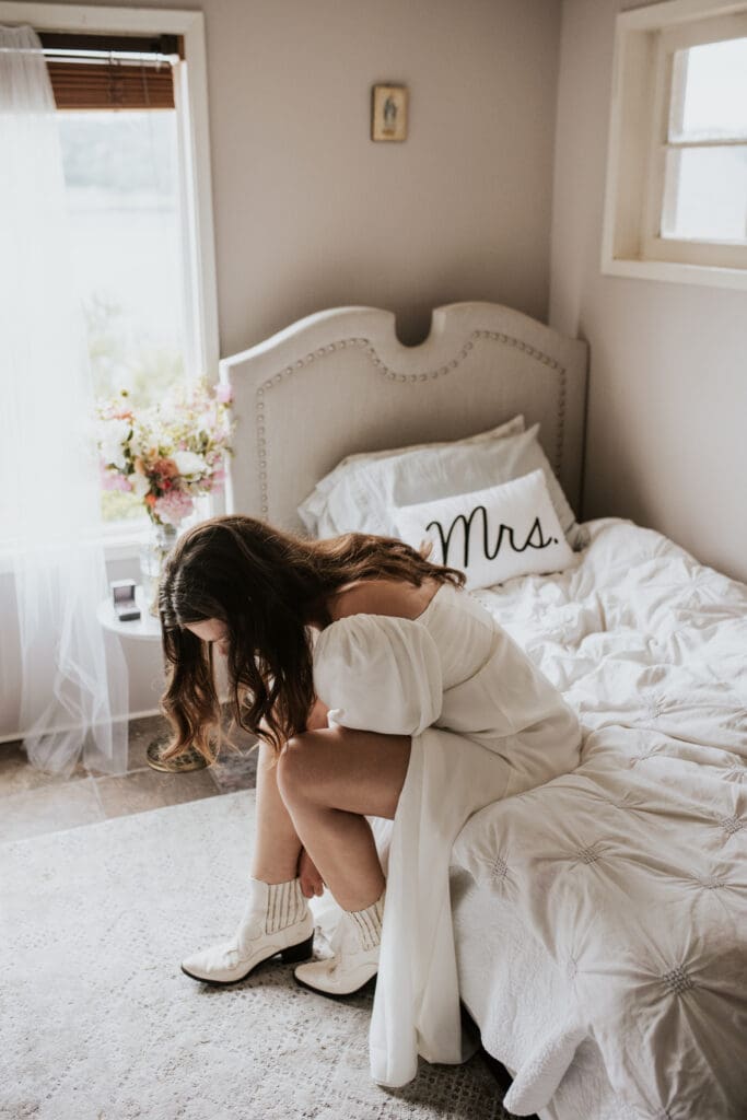 a woman putting on her white cowboy boot while sitting on a bed