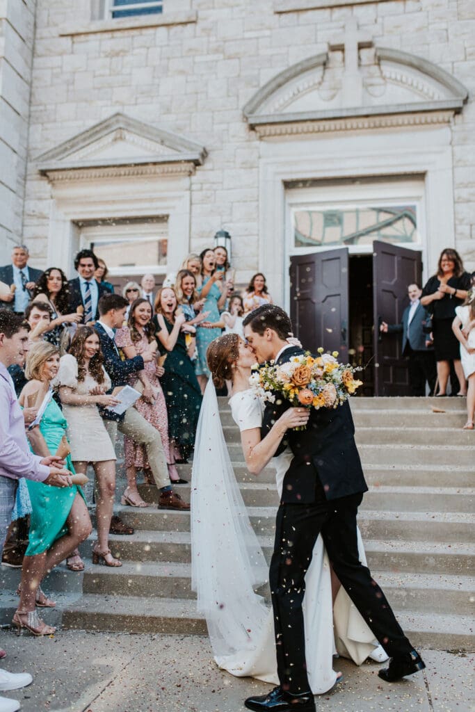 bride and groom kissing as guests throw flower petals at them in catholic ceremony exit
