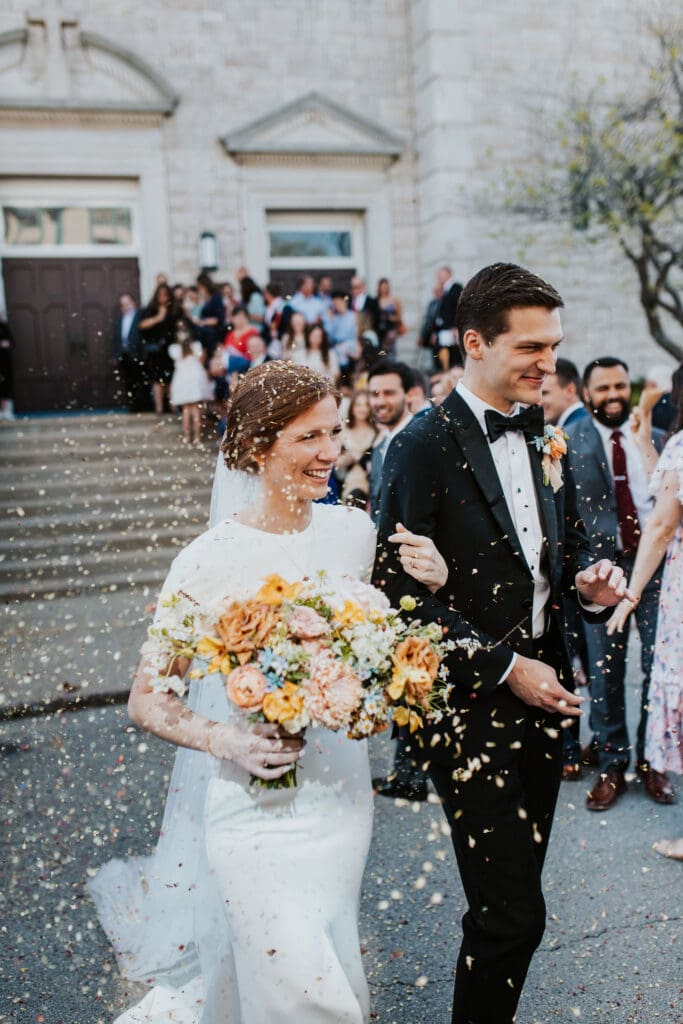 Bride and groom laughing as flower petals rain down outside the church doors