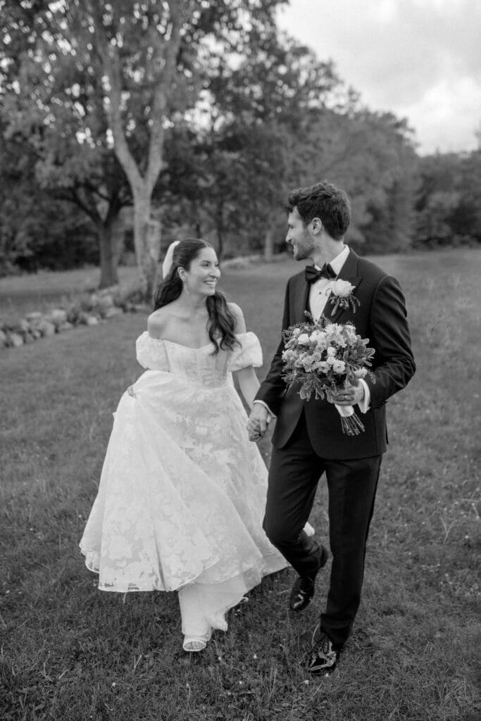 black and white photo of the bride and groom walking while smiling at each other and the groom is holding her bouquet