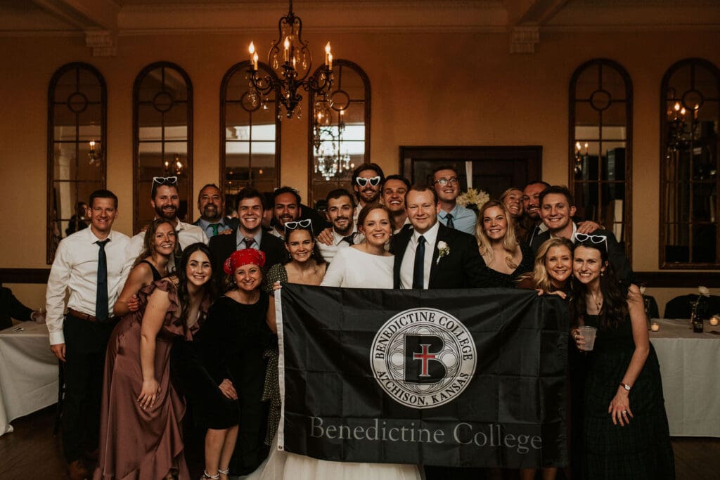 Bride and Groom stand with flag surrounded by fellow alumni from benendictine college at an atchison, KS wedding reception