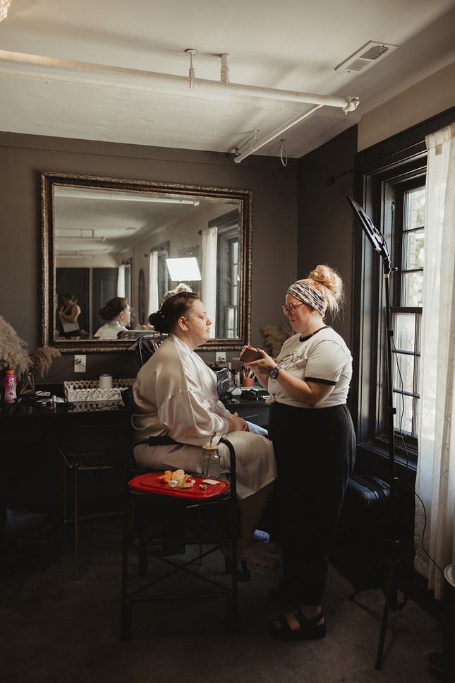 Bailey Rose Makeup Artistry applying bridal makeup in a cozy getting ready space with window light and mirror reflection