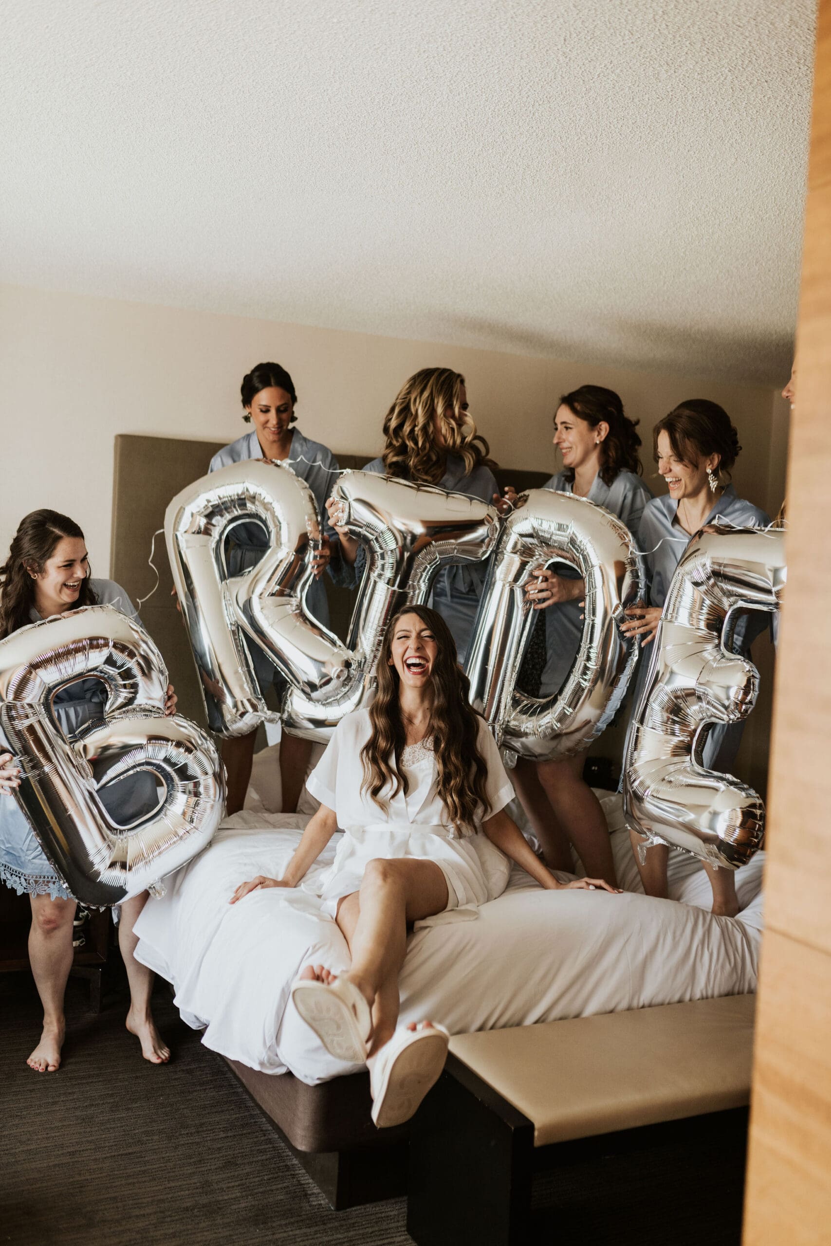 Bride celebrating with bridesmaids holding silver bride balloons during wedding morning.
