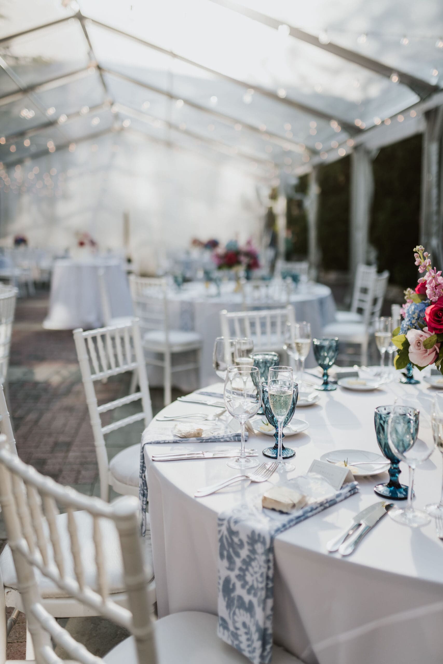 Outdoor wedding reception tables under clear tent with string lights and floral centerpieces.