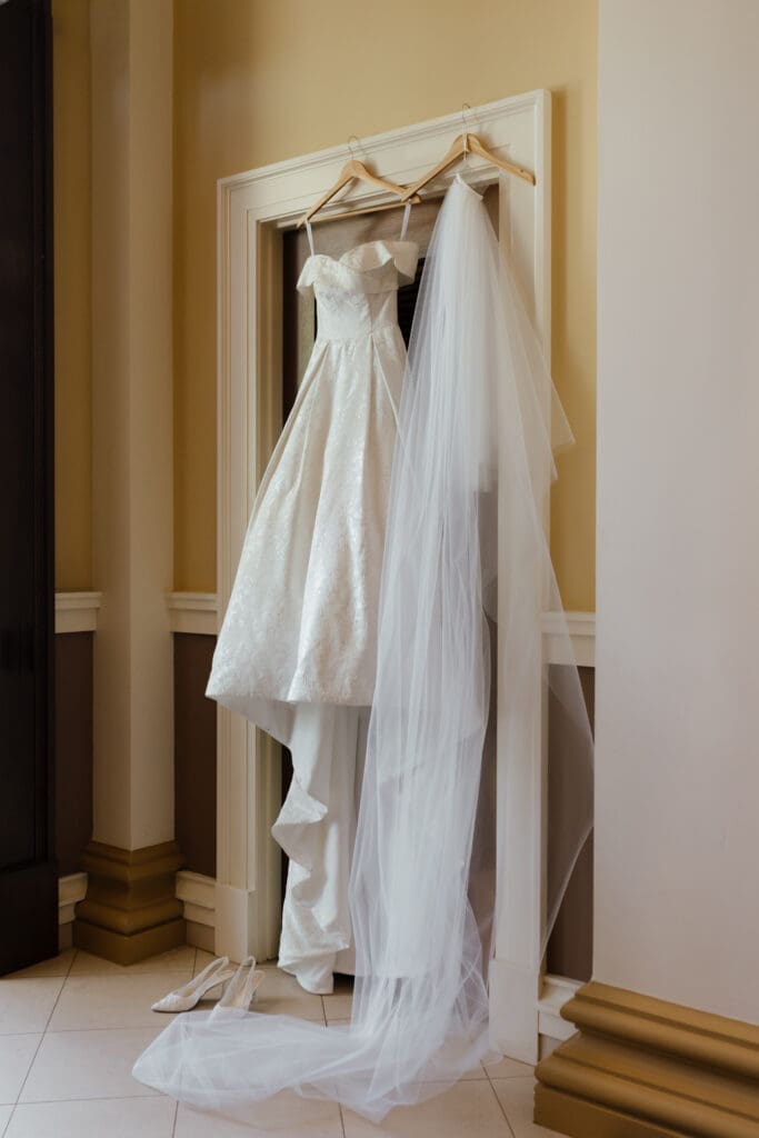 A detailed jacquard wedding ballgown with off-the-shoulder straps hangs in a doorway next to a long tulle veil on wooden hangers. White wedding shoes are on the floor below.