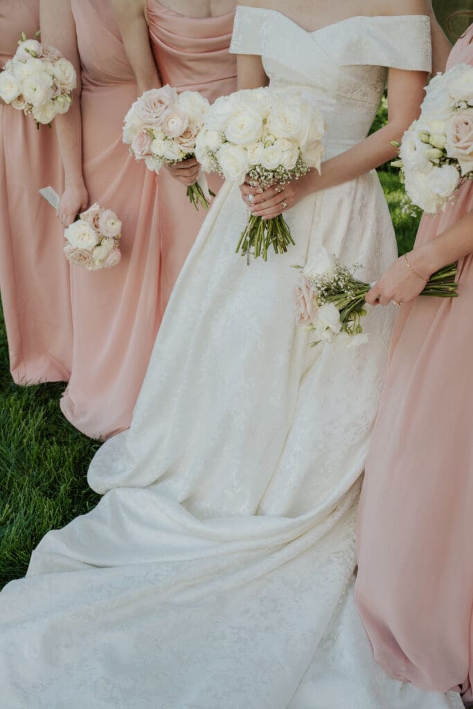 A close-up color photo of hands holding three distinct bouquets: one with white roses and greenery, one smaller with baby's breath, and one a white rose cluster. Bridesmaids in light pink dresses and the bride in her jacquard ballgown frame the shot against outdoor greenery.