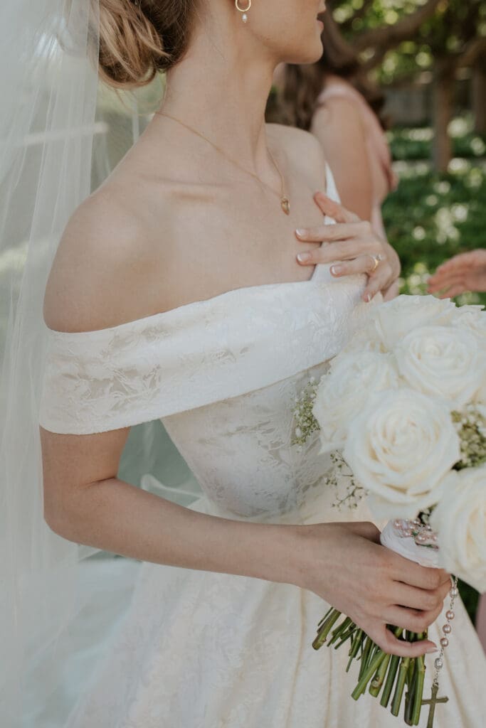 A detailed color close-up of the bride from the neck down, showcasing the fabric of her jacquard ballgown with off-the-shoulder straps, a simple gold necklace with a pendant, a pearl earring, and her hand holding a white rose bouquet adorned with a clear crystal rosary. A hand is adjusting the strap.