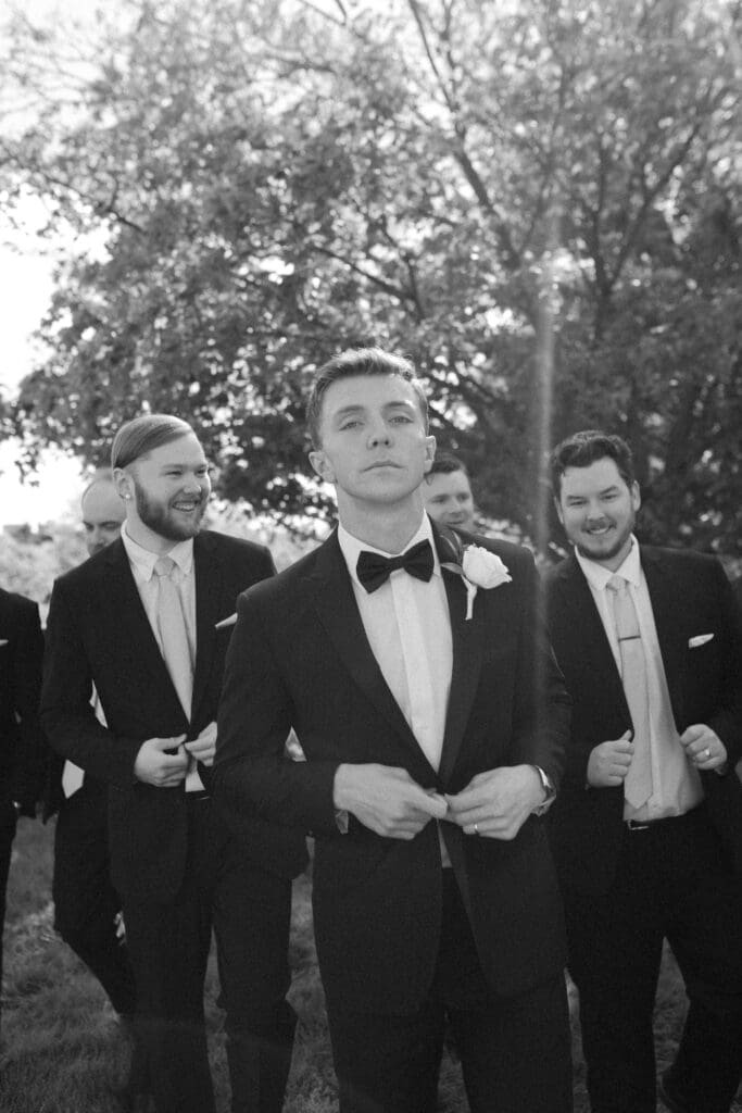A candid black and white photo of the groom and his groomsmen outdoors with trees. The groom is centered, adjusting his tuxedo jacket with both hands, looking pensive at the camera. Three groomsmen are behind him smiling and walking.