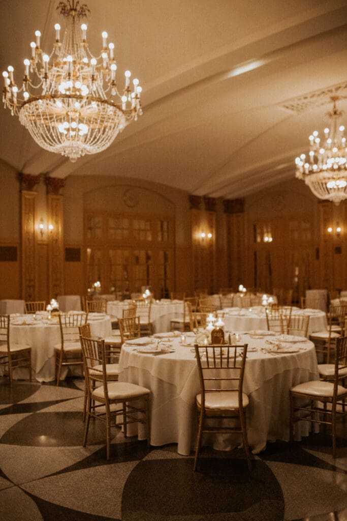 A wide-angle color photo of the Hilton President Grand Ballroom, empty before the reception. Features the large crystal chandeliers, golden walls, a central mirror, and the detailed pattern floor. Tables are set with white cloths, Chiavari chairs, and lit candles.