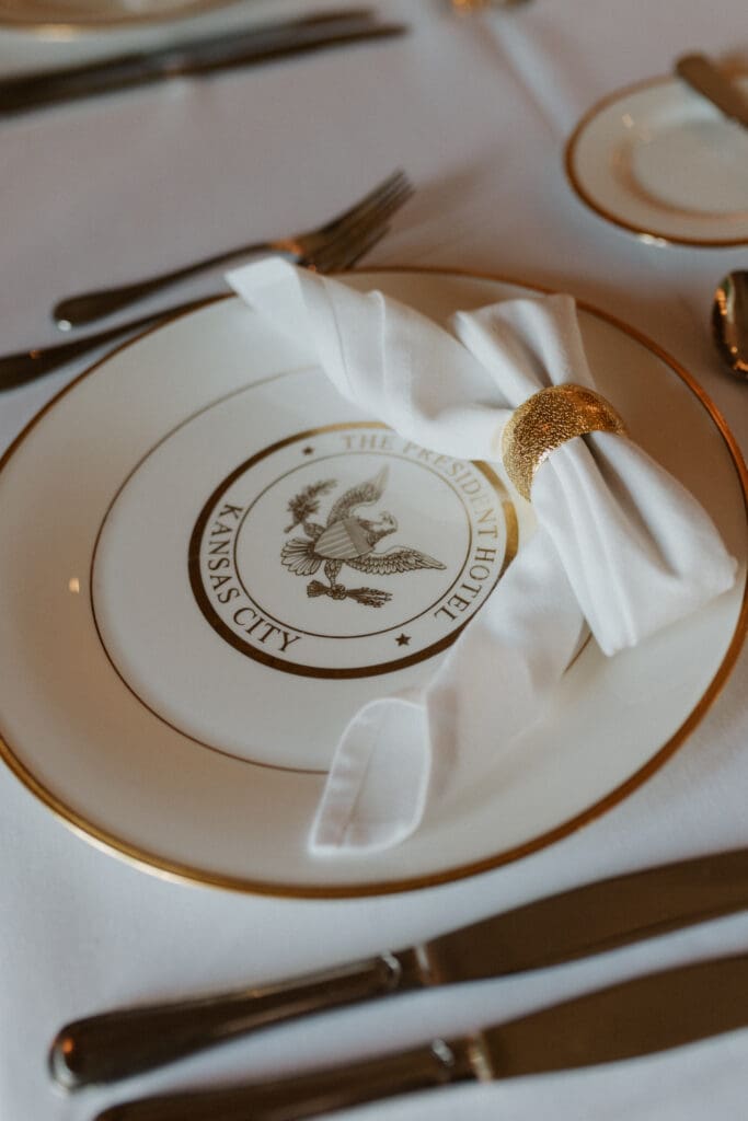A detailed close-up color photo of a formal place setting on a white tablecloth. A white and gold-rimmed plate is embossed with the official circular logo that reads "THE PRESIDENT HOTEL KANSAS CITY" with an eagle emblem. A golden napkin ring holds a white napkin. Cutlery is visible.