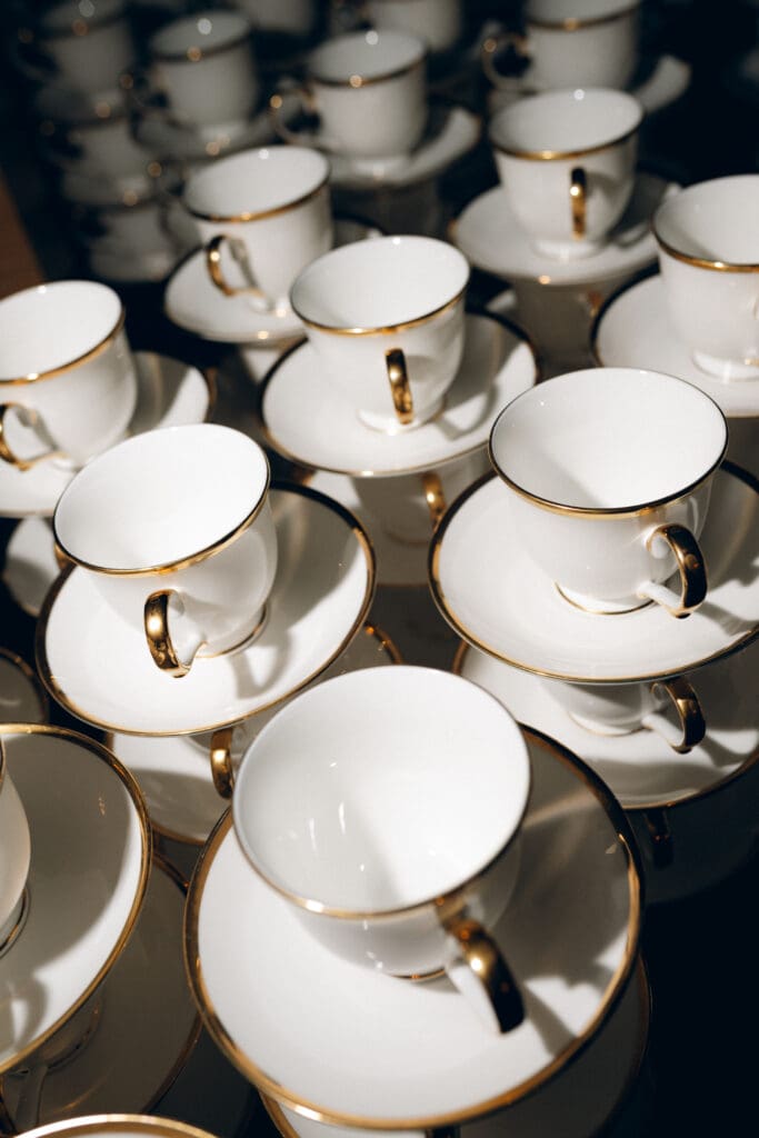 A detailed close-up color photo of multiple stacked white and gold-rimmed coffee cups and saucers, prepared in stacks, with warm light highlighting their elegant design and gilded handles.