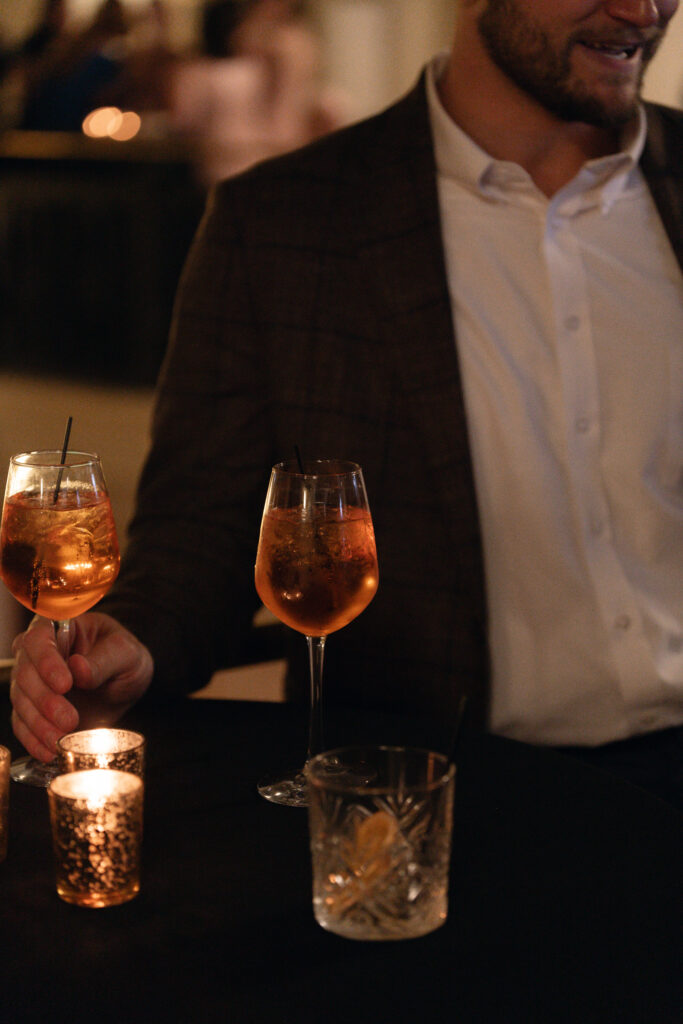 A detailed close-up color photo of the cocktail hour table at the Hilton President reception. Lit votive candles create a warm glow. A hand holds one large glass of an orange signature cocktail (Aperol Spritz). Another rocks glass with a large clear ice cube is in the foreground.
