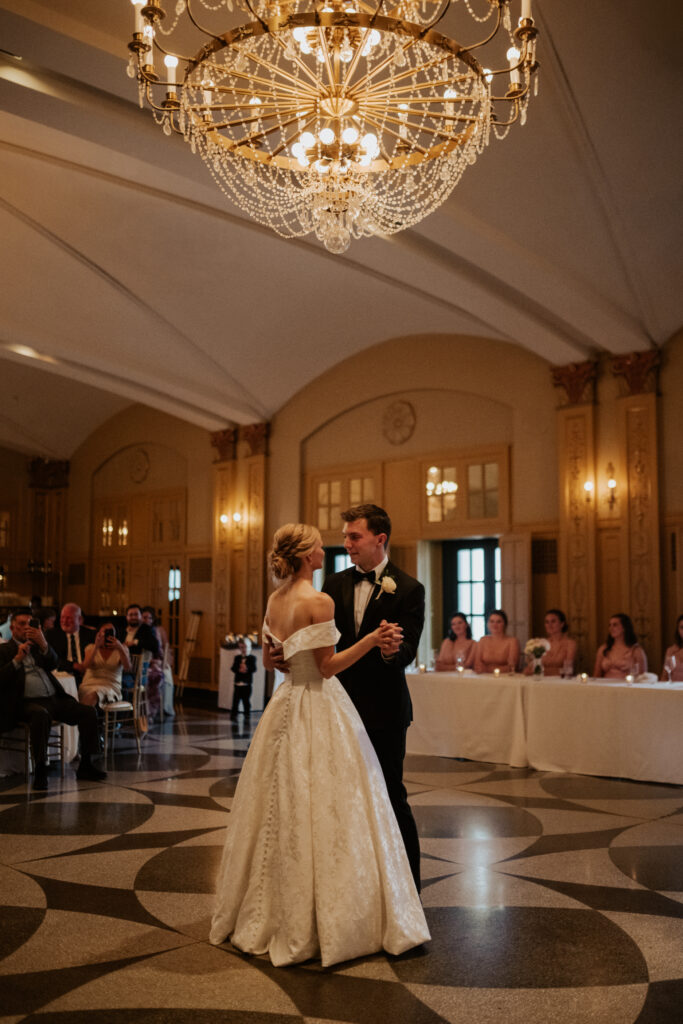 A wide-angle color photo of the bride and groom sharing their sweet first dance in the Hilton President Grand Ballroom. A massive crystal chandelier is overhead, and guests are seated at tables in the dark, warm ballroom background, many watching with warm smiles.