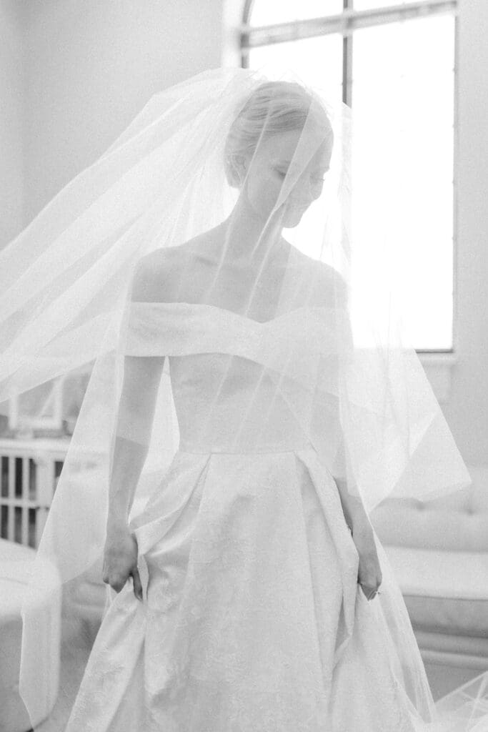 An artistic black and white wedding portrait of the bride looking down, her face pensive behind a flowing tulle veil that slightly obscures her smile.