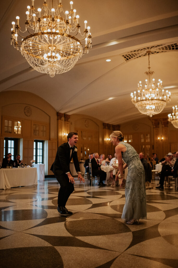 A dynamic color photo of the groom and his mother sharing a fun, choreographed first dance in the Hilton President ballroom. They are both smiling broadly. Guests at the seated tables are laughing and watching. A large chandelier is dominant overhead.