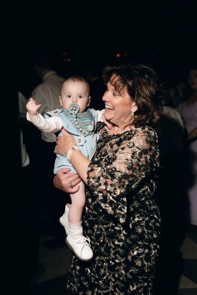 A high-energy direct flash color photograph taken during open dancing at the Hilton President Kansas City. An older woman (presumably a grandmother) is centered on the dance floor, smiling broadly while gently holding a baby who is wearing tiny noise-canceling headphones. Other wedding guests are blurred in motion around them.