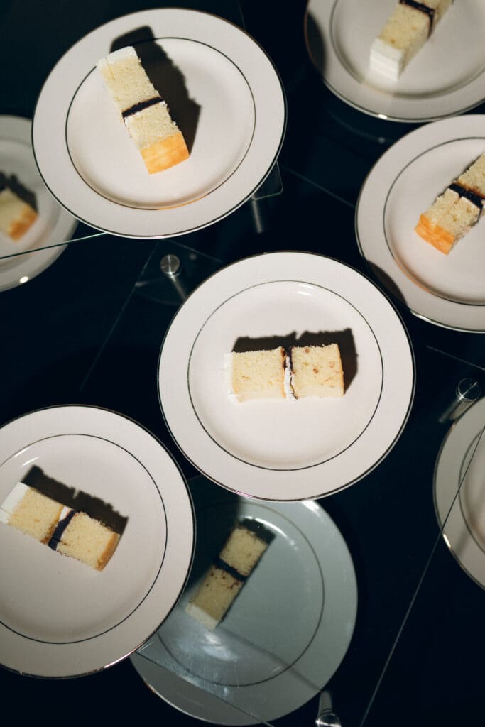 A close-up color photo of individual wedding cake slices plated on white and gold-rimmed Hilton President plates. Captured with direct flash, the lighting makes the white frosting pop against the dark reception background.