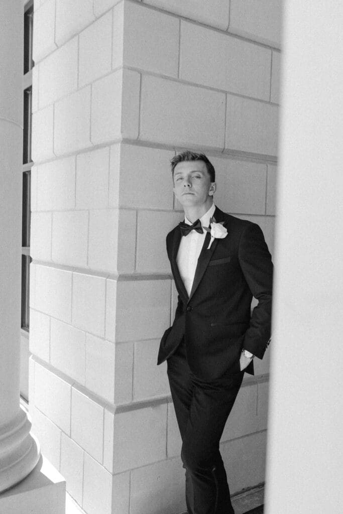 A confident black and white portrait of the groom in a tuxedo with a single rose boutonnière, leaning against the classic stone pillar architecture of St. Michael the Archangel church in Leawood.