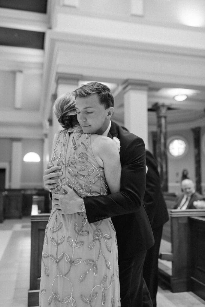 An emotional black and white candid photo of the groom hugging his mom wearing a sparkly dress, sharing a tender embrace during the wedding day celebration.