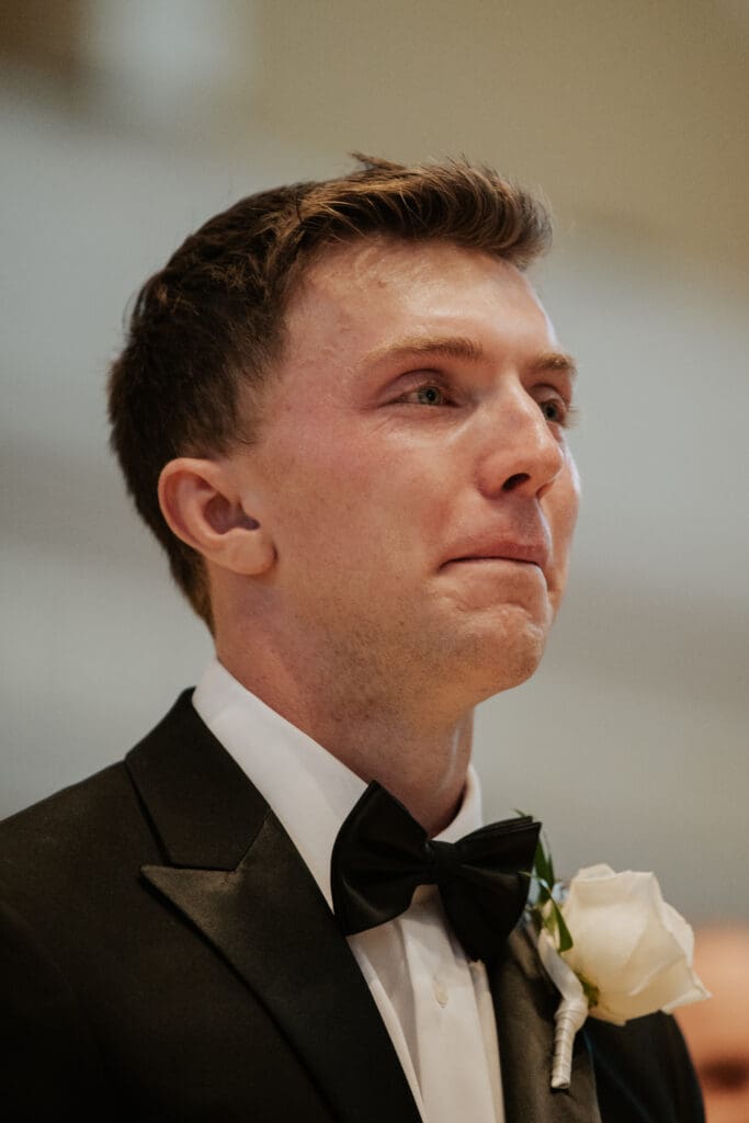 A tight color portrait of the groom at the altar, eyes glistening with tears and looking down the aisle, wearing a black bowtie and a white rose boutonnière.
