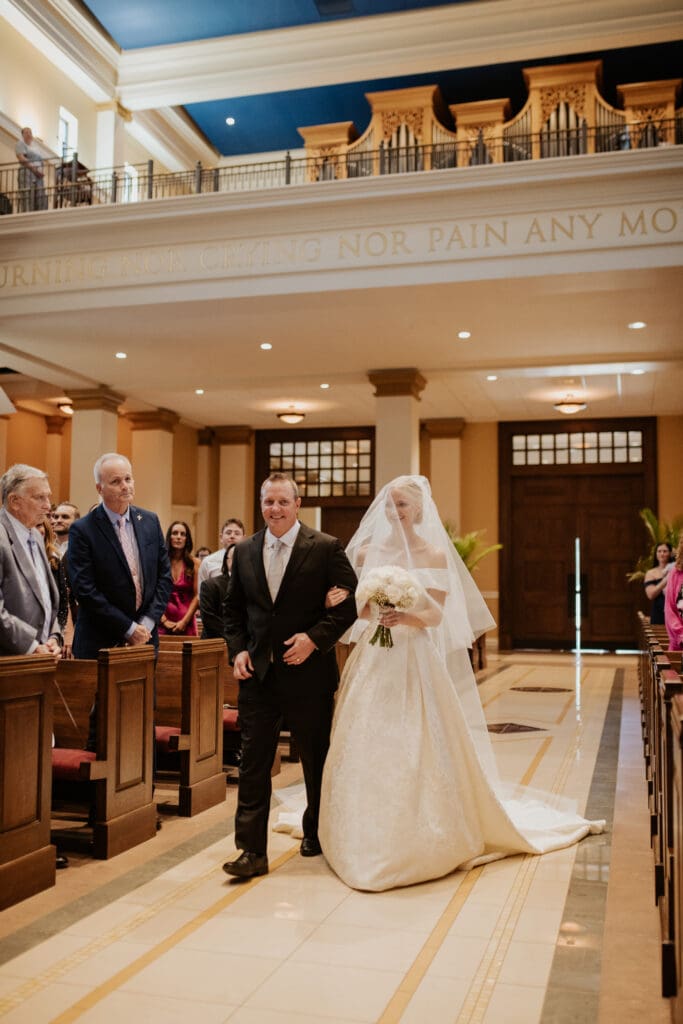 A wide view of the St. Michael the Archangel Leawood wedding processional, showing the bride’s father smiling as he walks her down the aisle. The grand pipe organ is visible above.