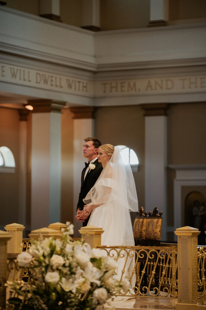 A side perspective of the bride and groom standing together at the altar of St. Michael the Archangel parish, with white floral arrangements and ornate white railings in the foreground. Golden text is visible on the wall above.