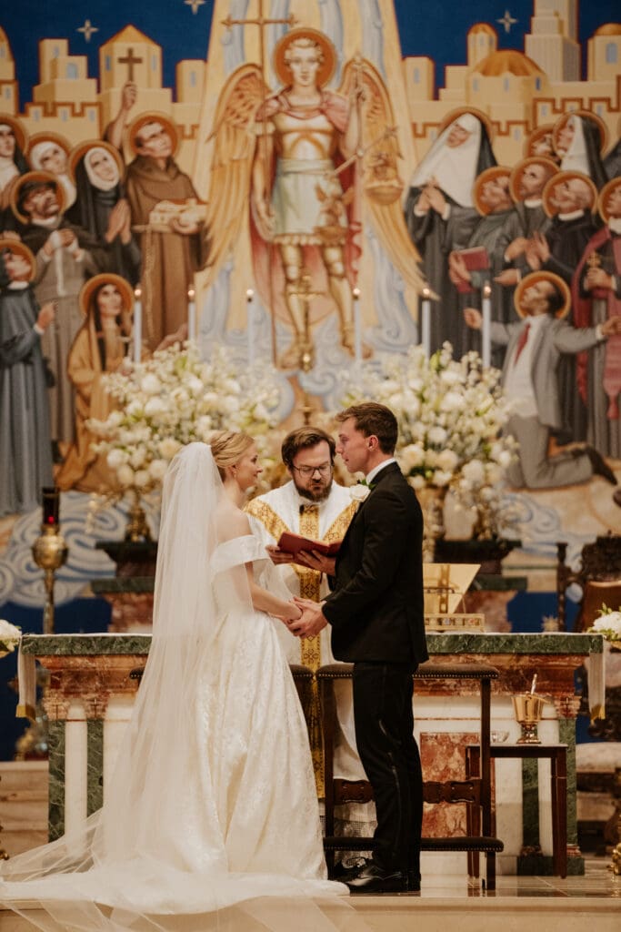 A detailed close shot of the bride and groom holding hands and exchanging vows at the altar of St. Michael the Archangel, Leawood. The colorful mosaic of St. Michael is behind them as a priest observes.