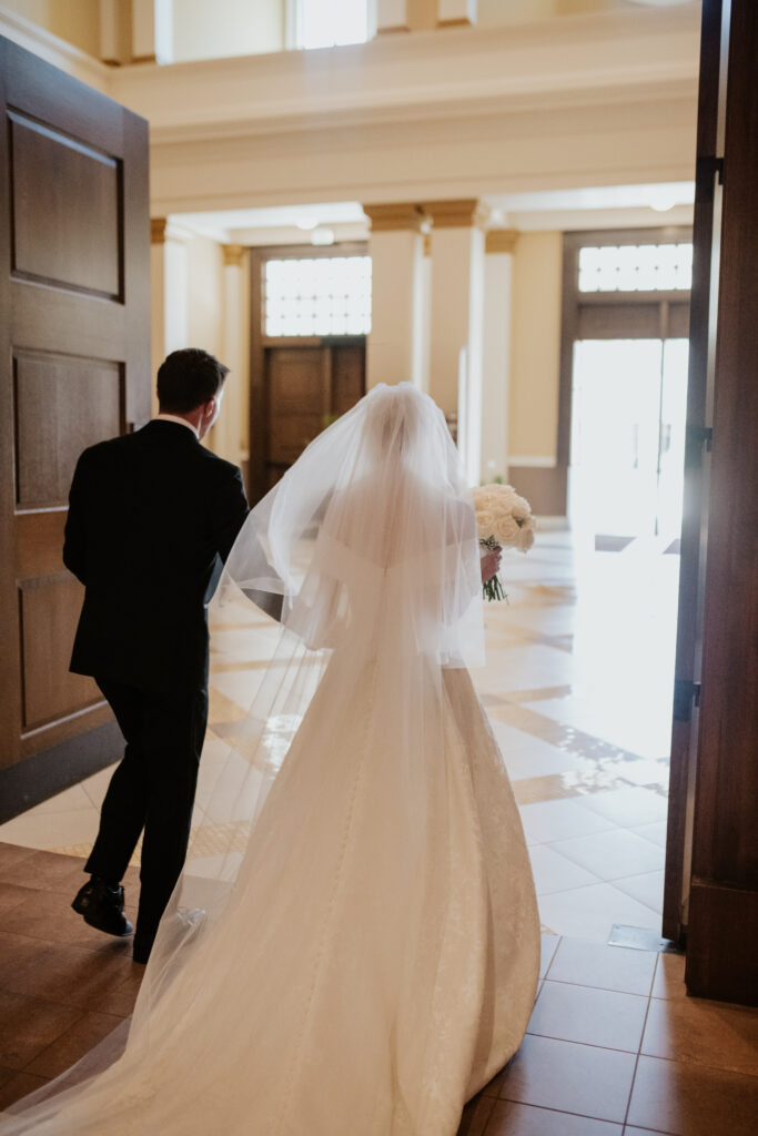 View from behind of the newlyweds hand in hand, walking away from the altar and toward the exit doors of the church vestibule, natural light flooding the entryway.