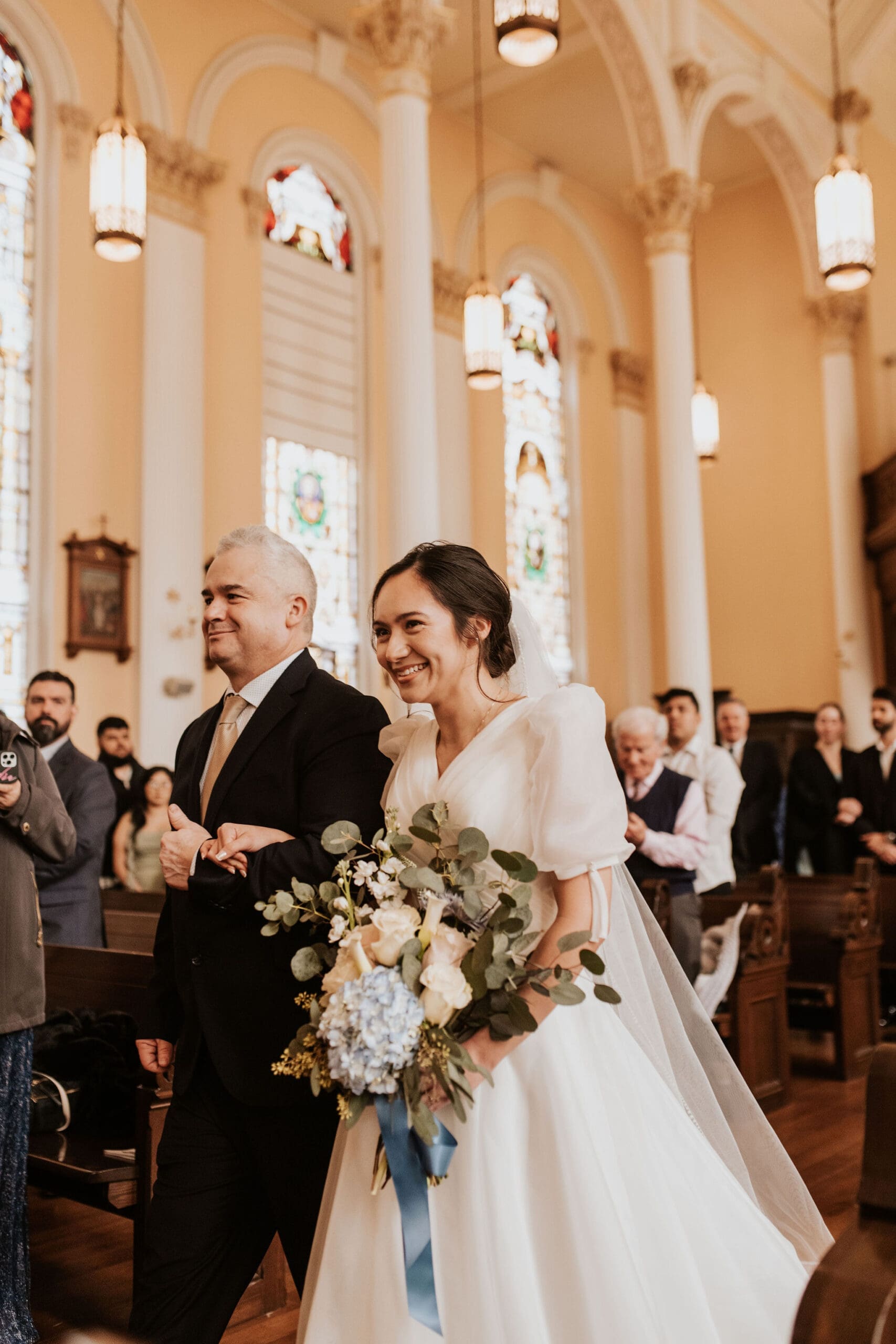 Emotional Catholic wedding processional with bride walking down the aisle beside her father in a historic church