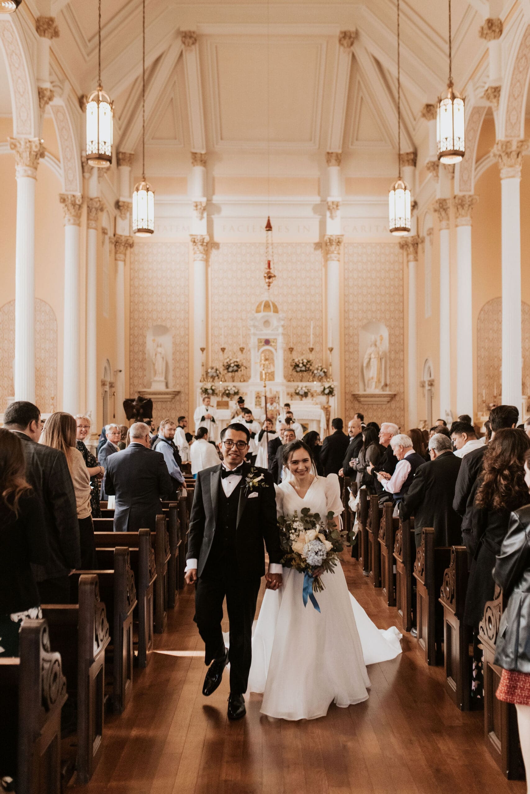 Newly married couple smiling during their recessional inside a softly lit Catholic church wedding ceremony