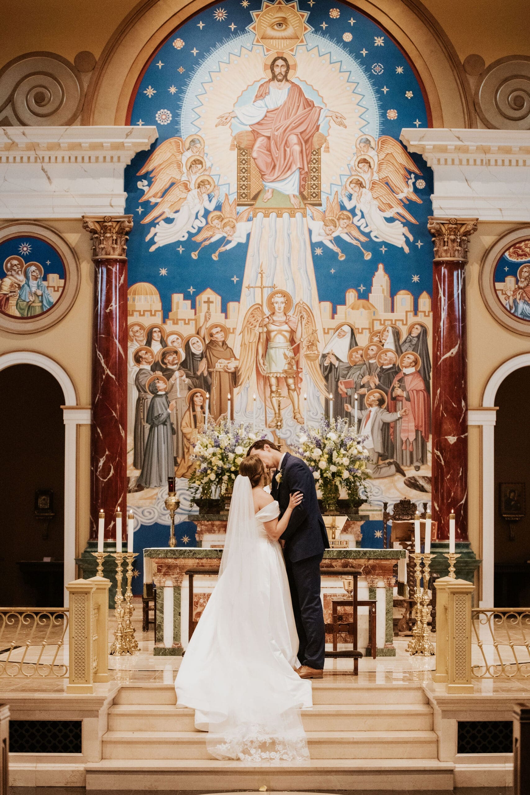 Bride and groom share a kiss at the altar beneath colorful religious artwork during a traditional Catholic wedding ceremony.