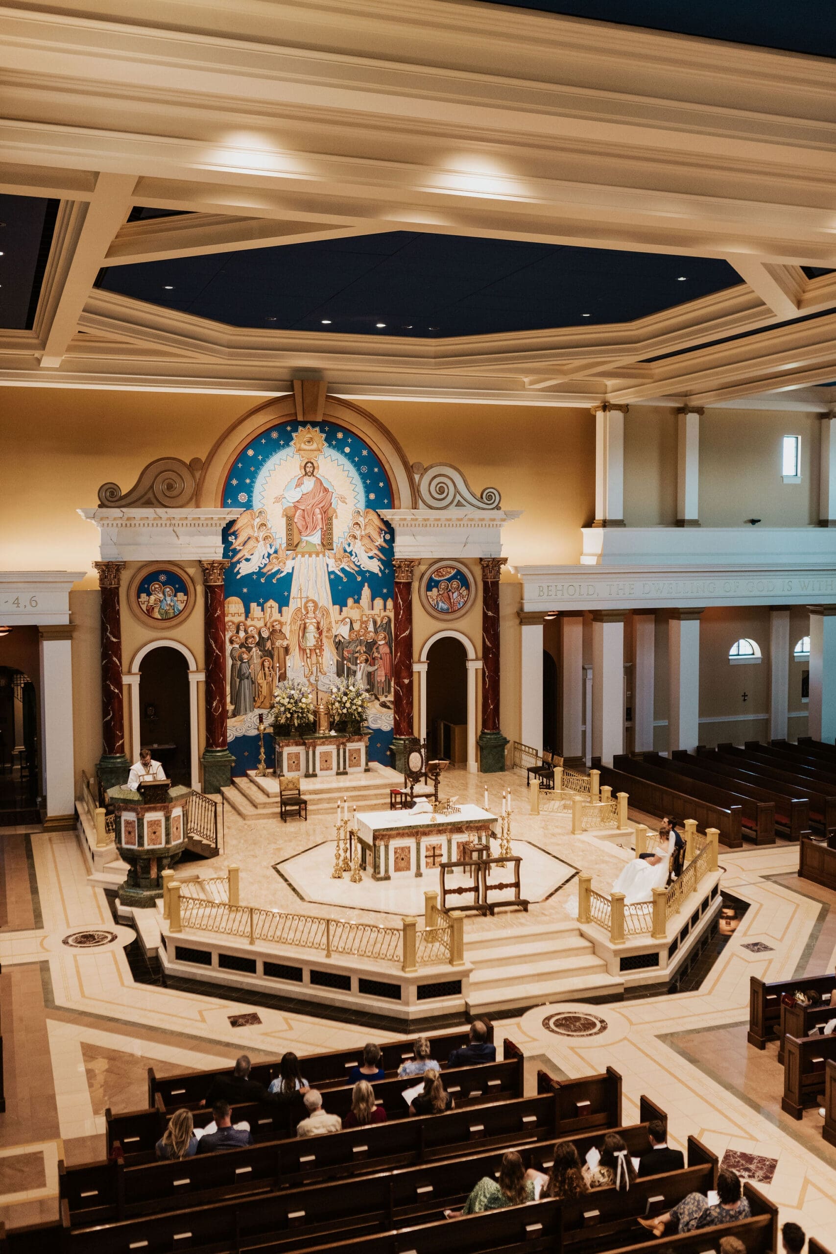 Wide view of a grand Catholic church ceremony with guests seated in wooden pews and the couple exchanging vows at the altar.