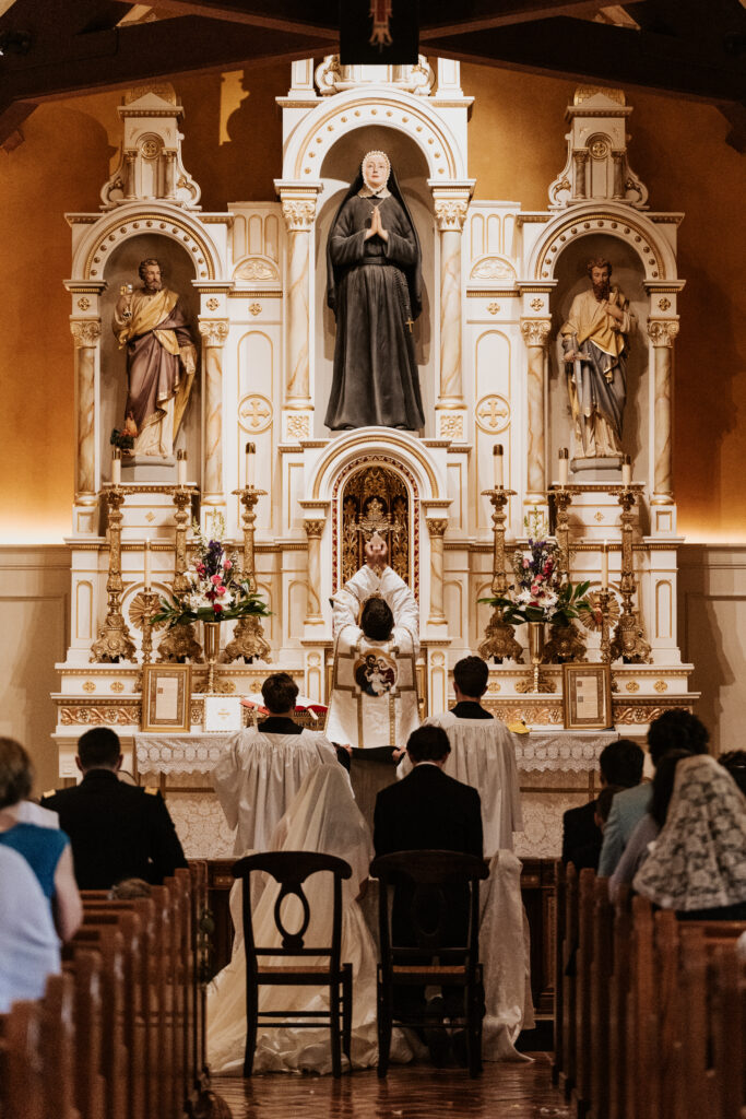 Traditional Catholic Latin Mass wedding ceremony at St. Rose Philippine Duchesne with the priest elevating the Eucharist at the high altar before the kneeling bride and groom.