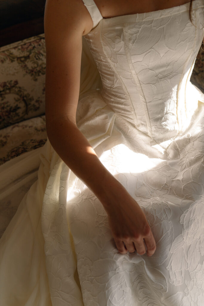 Detail shot of dappled afternoon sunlight catching the rich texture and structured bodice of a modern jacquard bridal gown inside an upstairs bedroom at Longview Mansion.