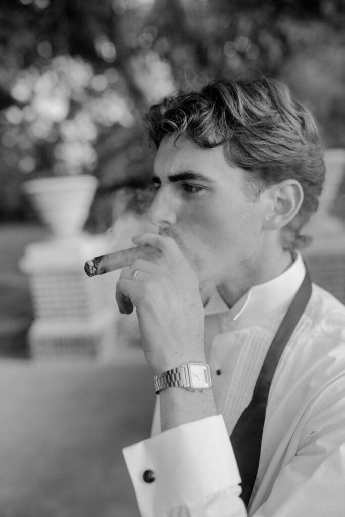 Black and white candid wedding portrait of the groom smoking a cigar outdoors at Longview Mansion, showcasing his new wedding ring.