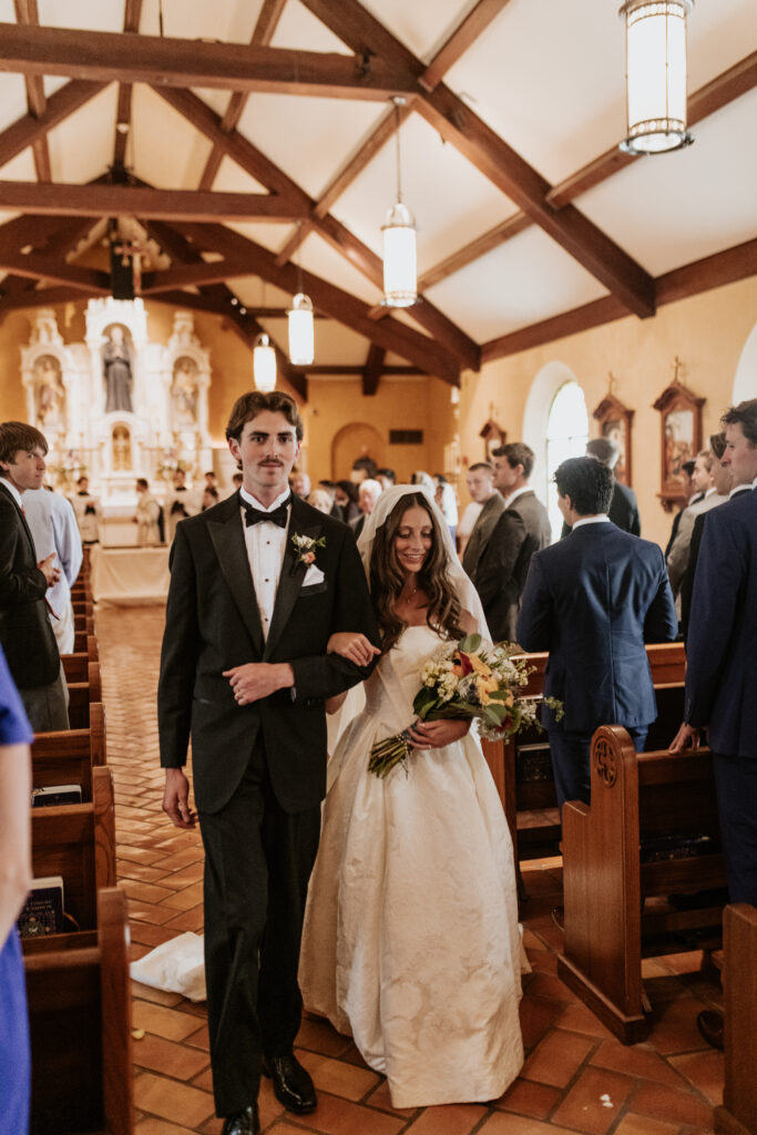 Newlyweds smiling during their wedding recessional down the brick-tiled aisle of St. Rose Philippine Duchesne Catholic Church under a vaulted timber ceiling.
