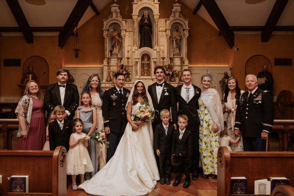 Large formal wedding family portrait at the altar of St. Rose Philippine Duchesne Catholic Church highlighting the bride's textured gown and guests in military uniform.