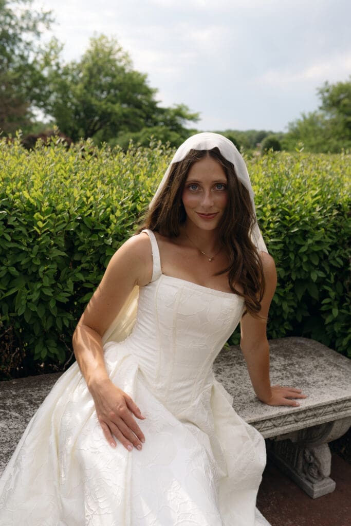 Close-up bridal portrait of a bride wearing a lace mantilla veil and a structured jacquard wedding dress sitting on a vintage stone bench at Longview Mansion.