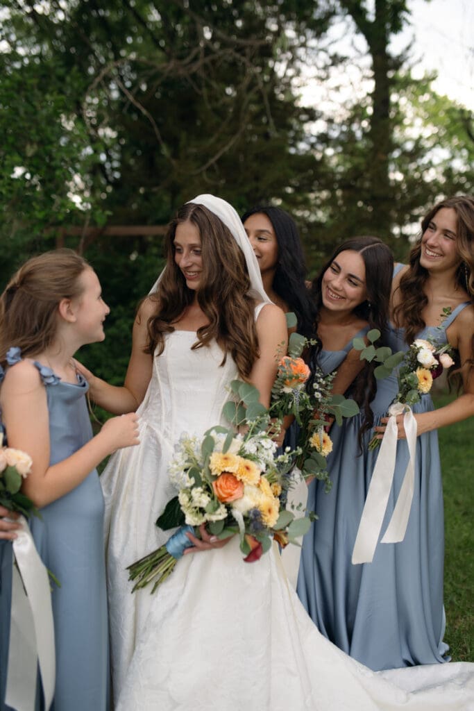 Bride looking down smiling at a young flower girl while bridesmaids in soft blue dresses hold organic summer bouquets on the lawns of Longview Mansion.