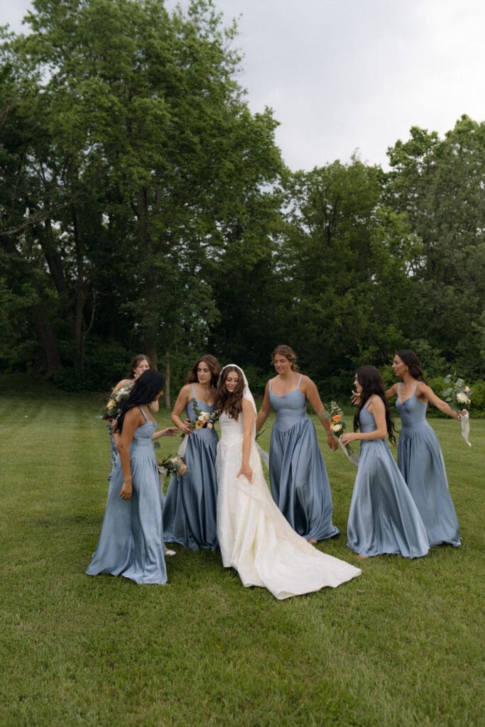 Editorial style wedding photography of a bride holding her train on the expansive green grass of Longview Mansion, surrounded by bridesmaids in matching slate blue gowns.