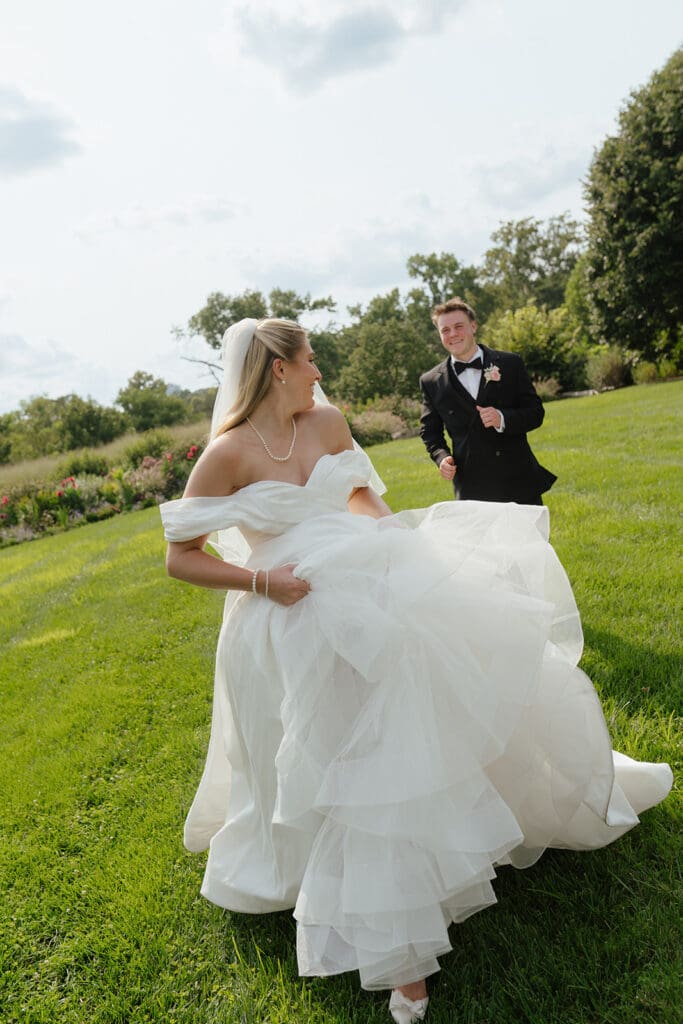 Bride laughing and holding her ballgown skirt while walking across a grassy lawn, groom smiling and jogging behind her