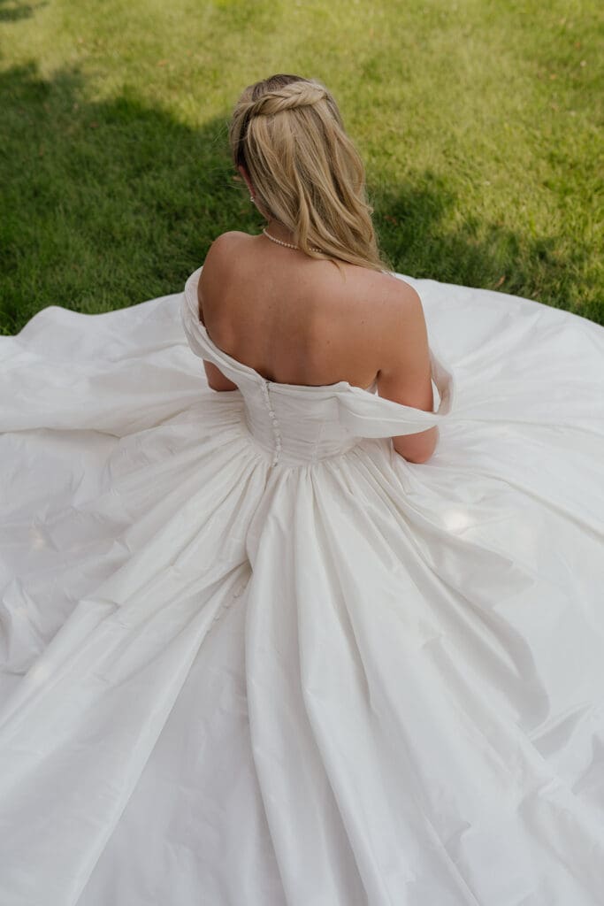 Overhead view of a bride's full ballgown skirt spread across the grass, seen from behind with braided half-up hair.
