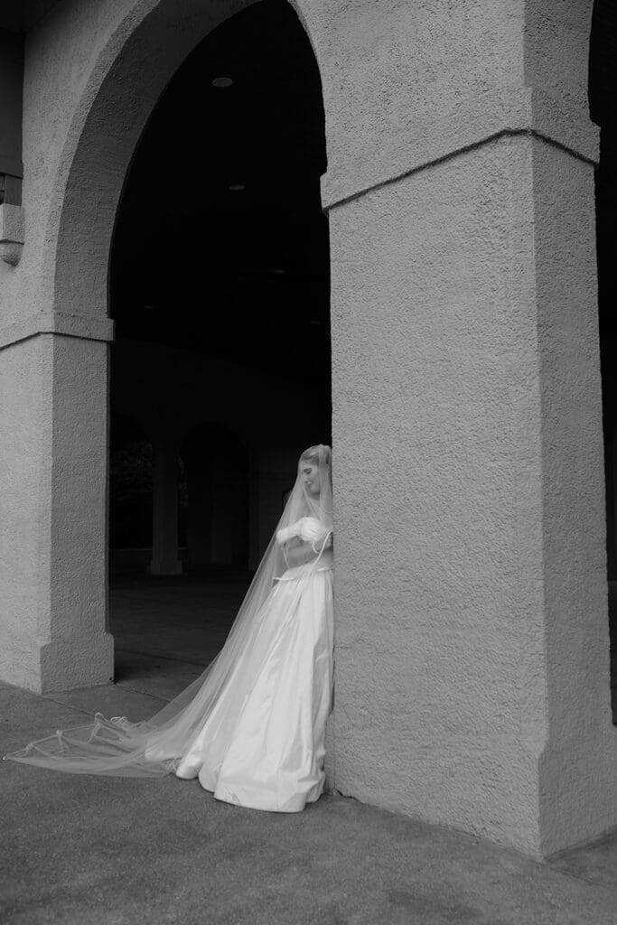 Black-and-white photo of a bride leaning against a stone archway pillar, veil trailing along the ground.