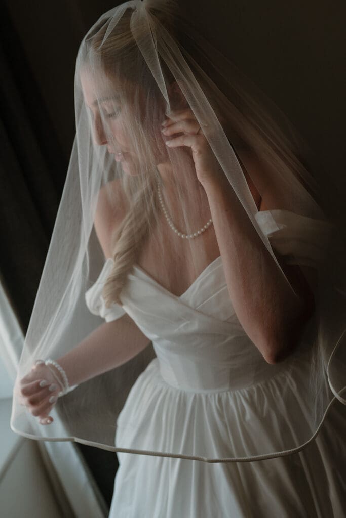 Bride in an off-the-shoulder wedding gown adjusting her veil beside a window, pearl necklace and bracelet visible through sheer tulle