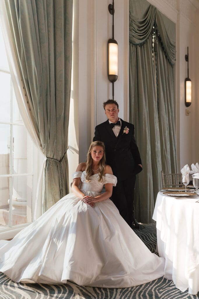 Bride seated in her ballgown with the groom standing behind her in a decorated ballroom in the St. Louis Marriott Grand with sage green drapery and table settings.