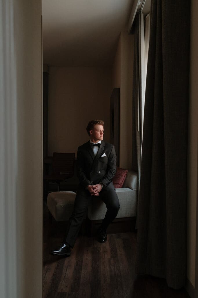 Groom in a double-breasted black tuxedo sitting on a bench by a curtained window, looking outside.