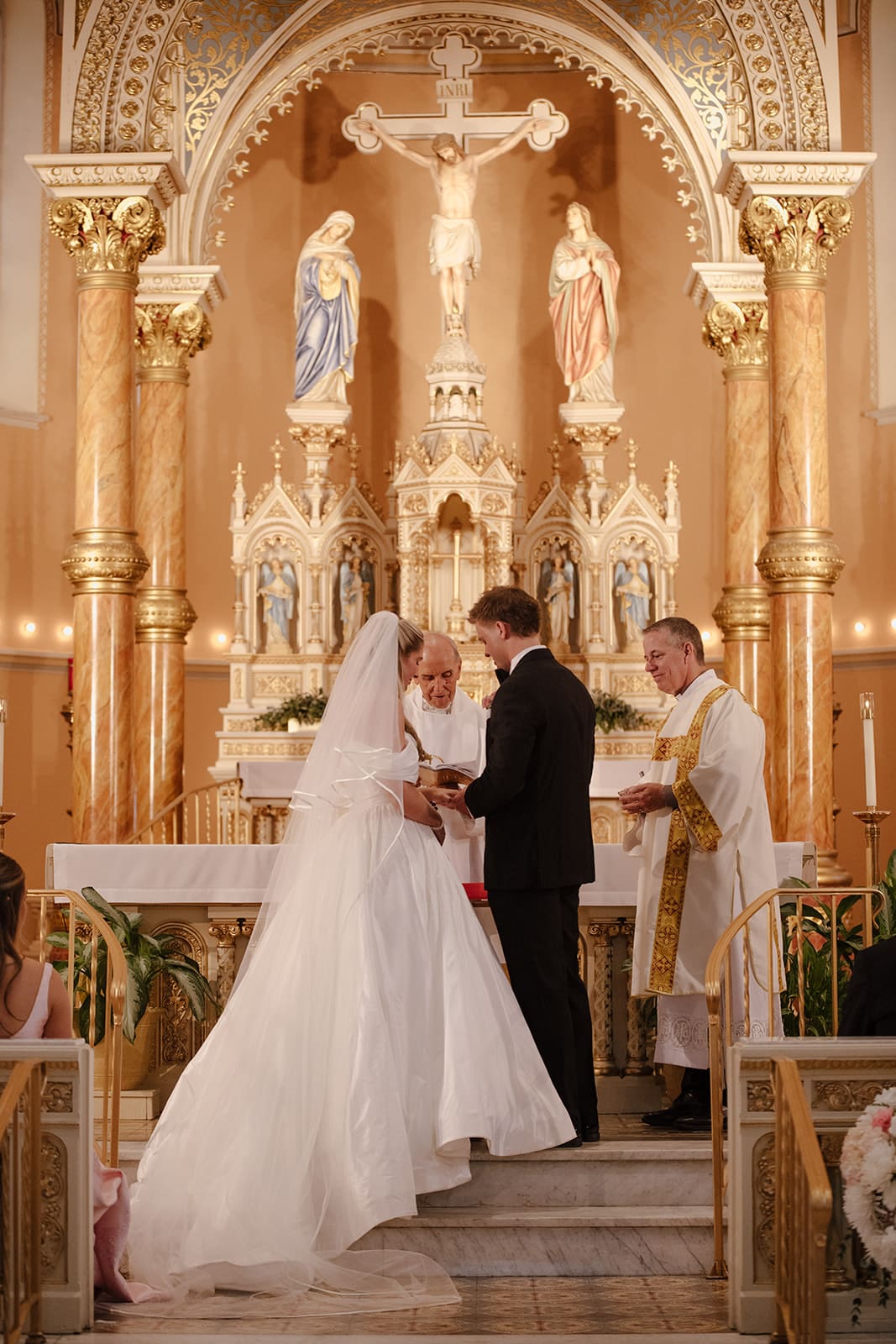 Bride and groom holding hands at the altar during their catholic wedding ceremony, with priests and an ornate gold altarpiece behind them.