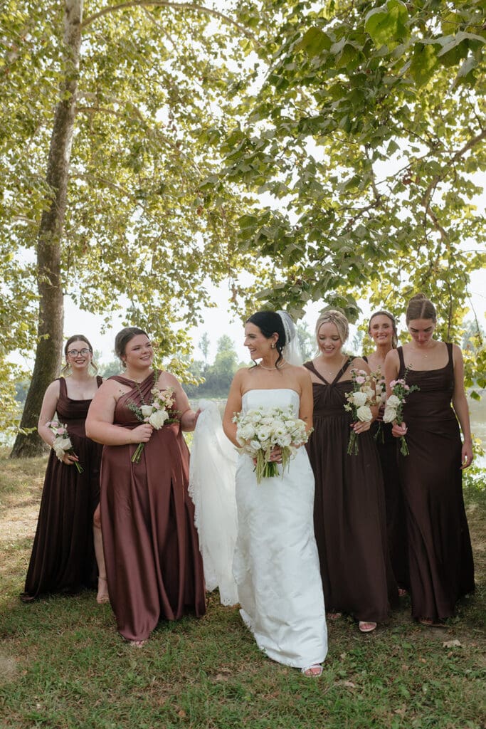 Bride walking with bridesmaids in brown gowns under trees at the Remington Nature Center