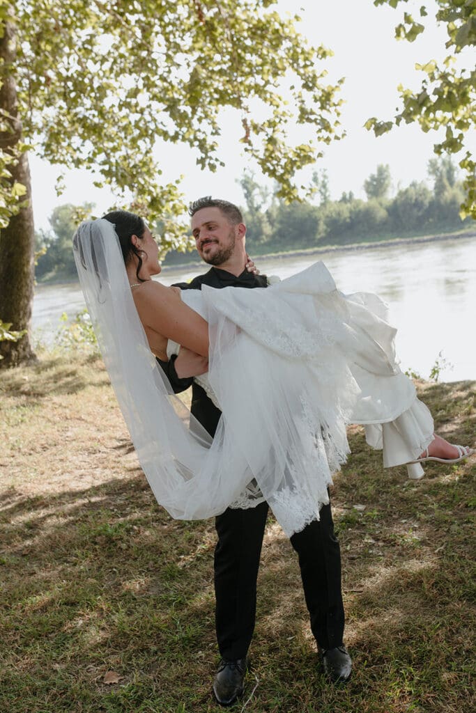 Groom carrying his bride near the river after their wedding ceremony