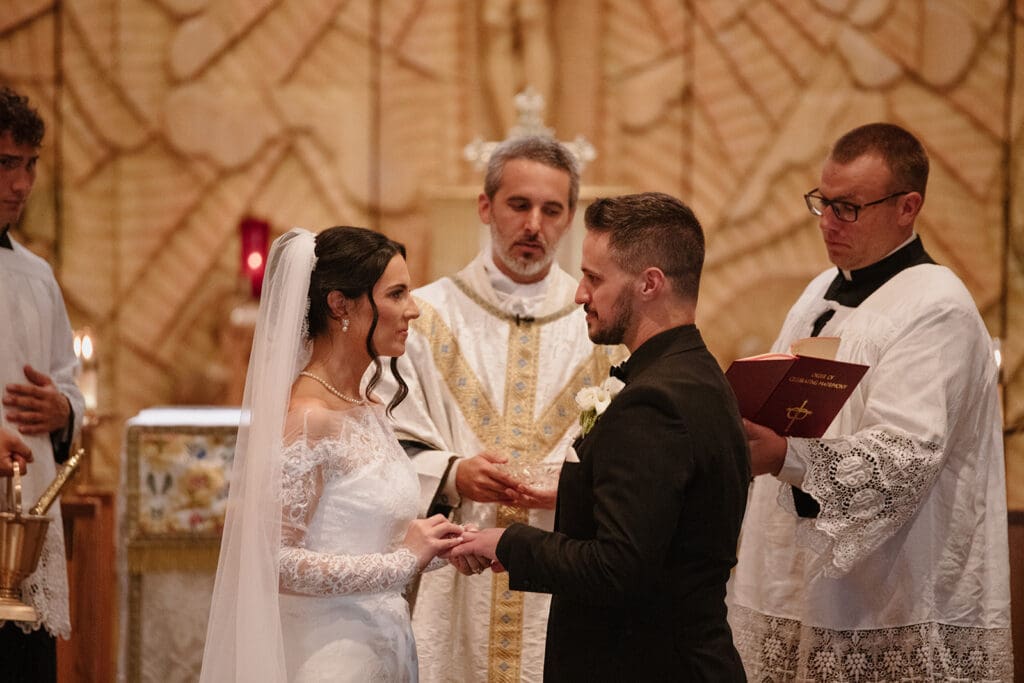 Bride and groom exchanging wedding rings during their ceremony at Our Lady of Guadalupe Catholic Church"
Title: "Exchanging rings at the altar